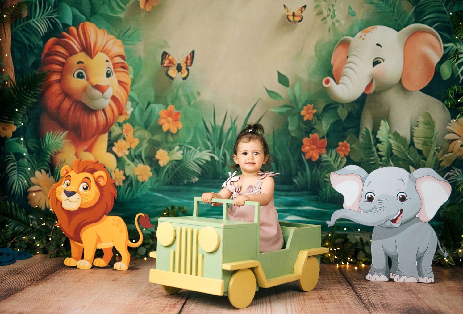 Toddler girl sits in toy jeep surrounded by jungle safari backdrop with cartoon lions and elephants for birthday photo.