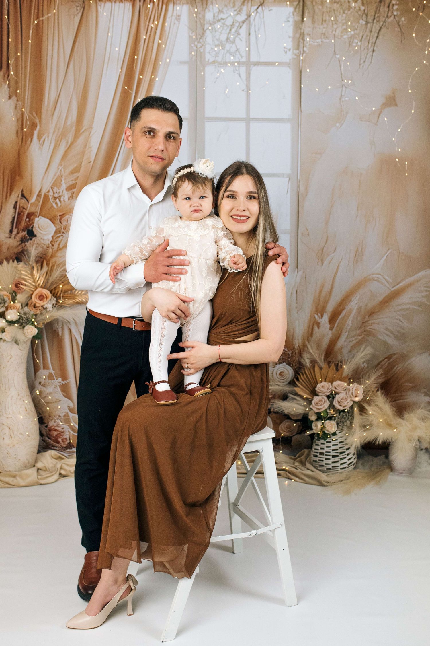 Family portrait with father, mother in brown dress, and baby girl posing in a decorated studio setting.