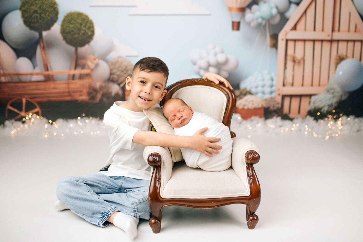 Older boy in white shirt and jeans poses with sleeping newborn in mini chair against pastel balloon backdrop.