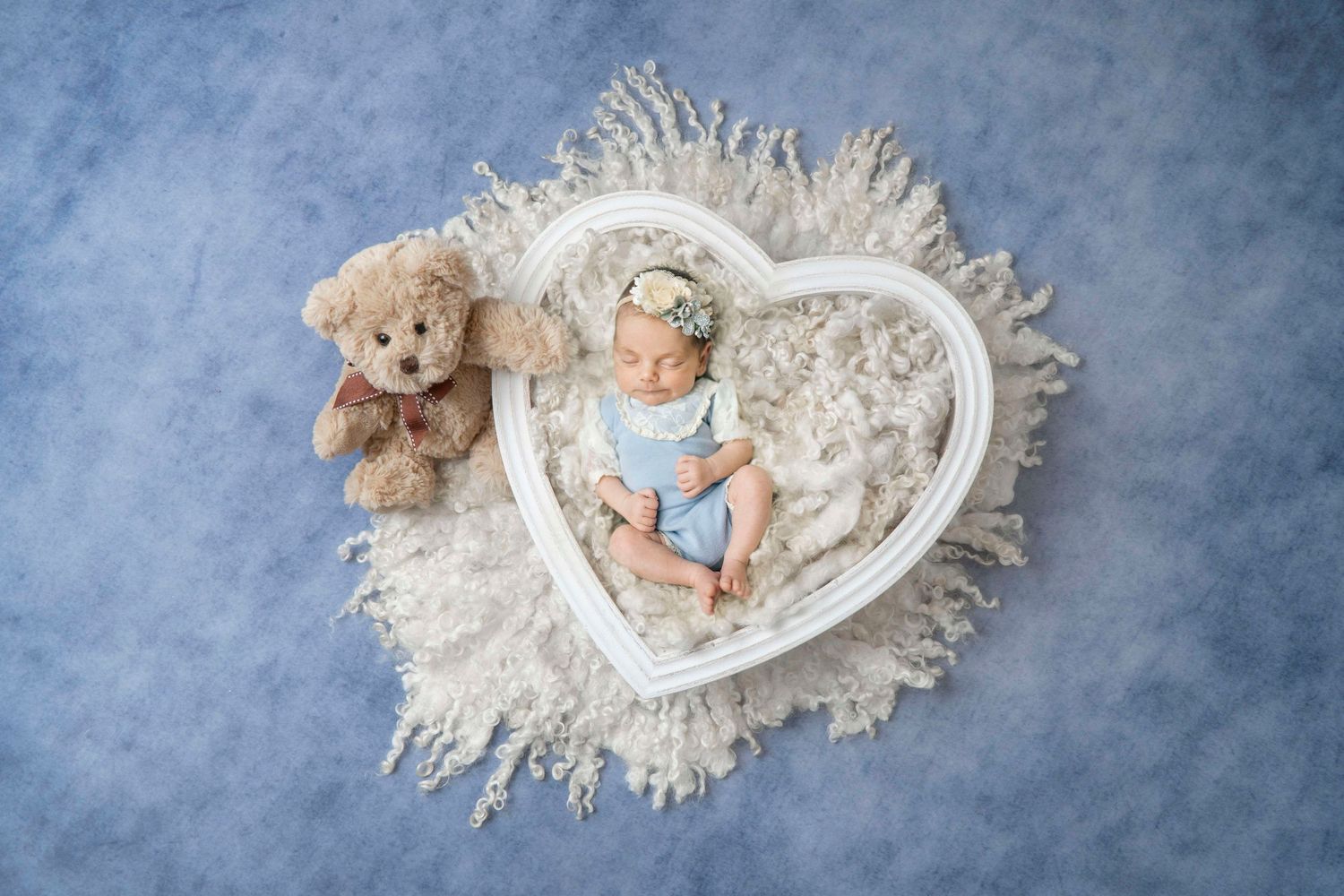 Newborn baby in blue romper sleeping in white heart-shaped bowl with teddy bear on blue background.