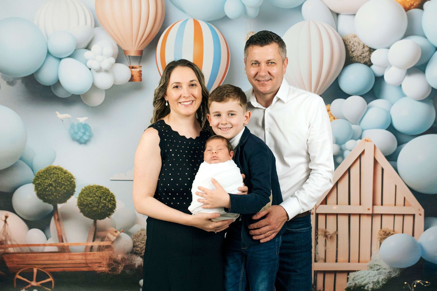 Family of four posing in front of a pastel balloon backdrop with hot air balloons, parents holding newborn baby.