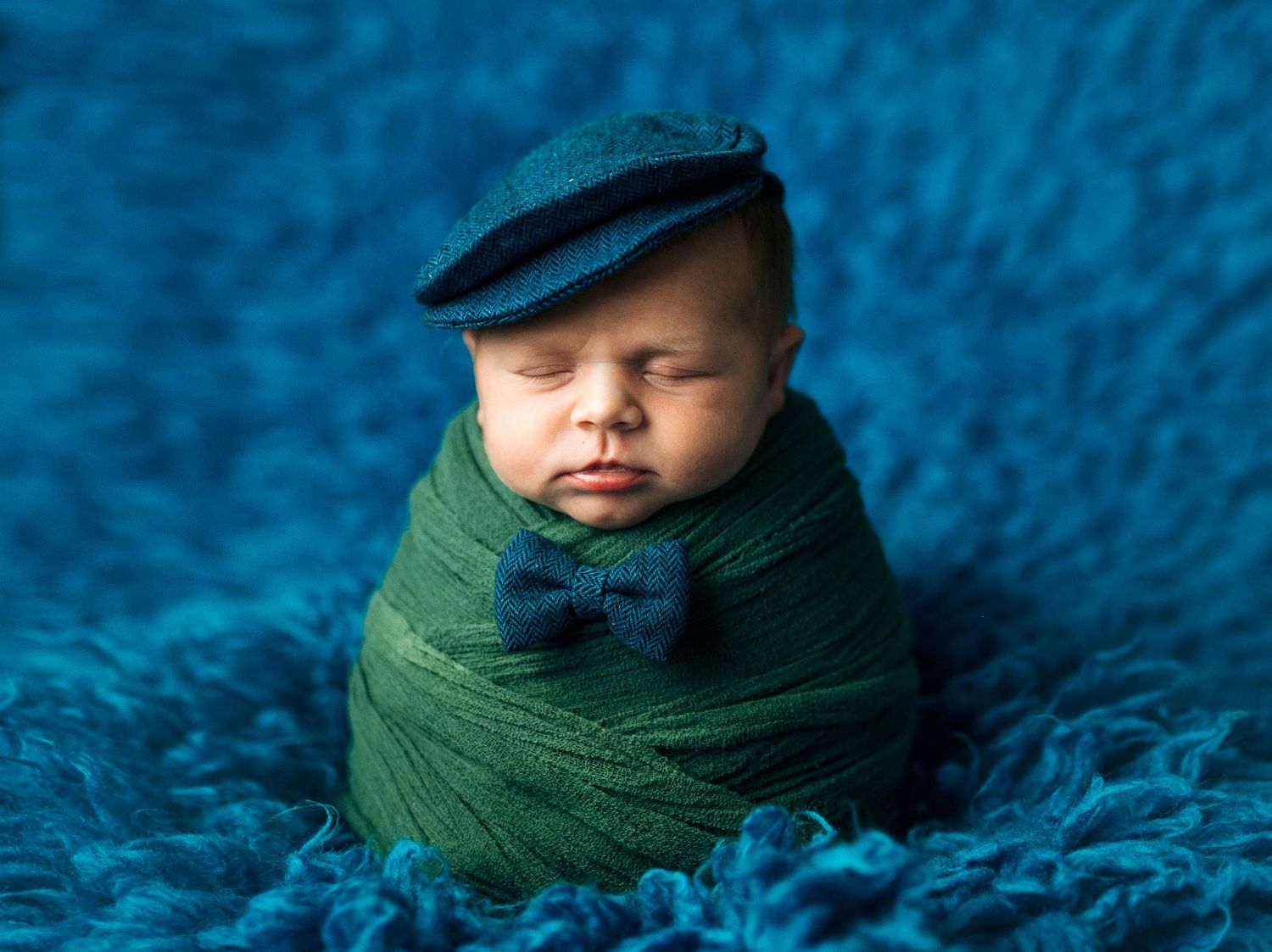 Newborn baby wrapped in green swaddle wearing blue flat cap and bow tie on teal fluffy background.