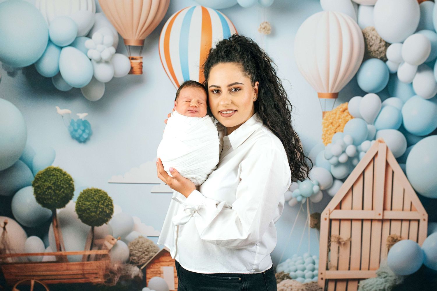 A smiling woman holds a swaddled newborn baby against a festive backdrop of blue balloons and hot air balloons.