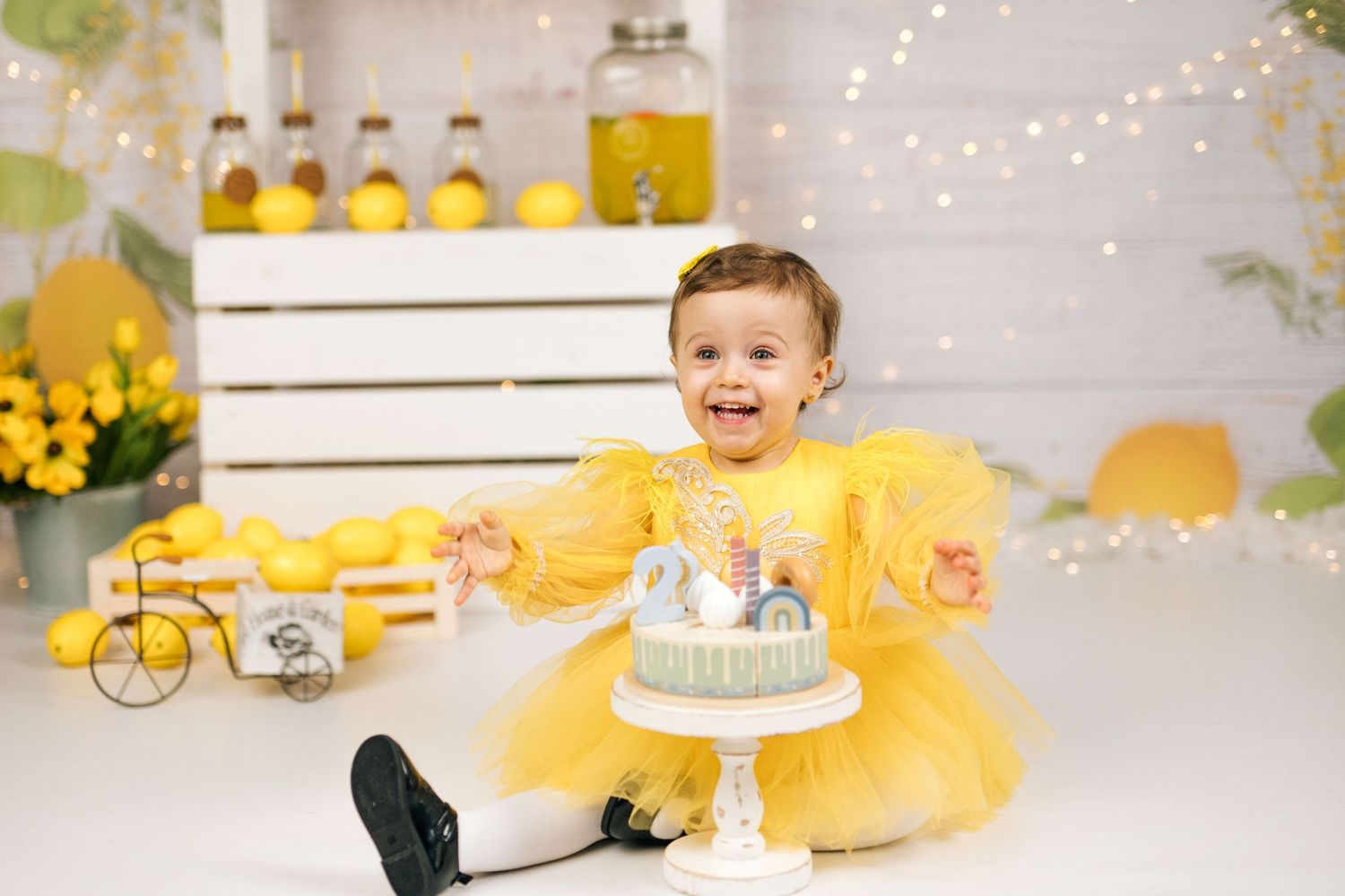 Smiling toddler girl in yellow tutu dress sits beside a small birthday cake in a yellow-themed party setting.