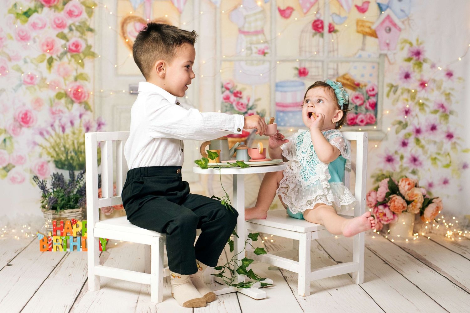 Two young children share a tea party at a small white table with a floral backdrop and birthday decorations.