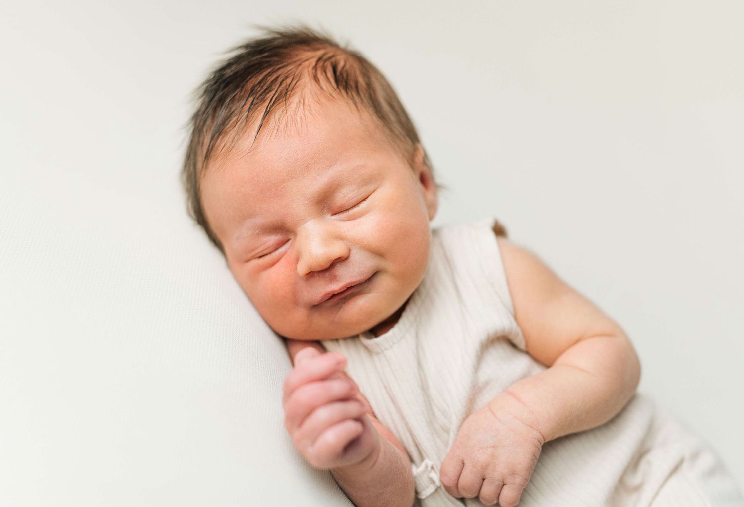 Peaceful newborn baby smiling in white onesie, lying on soft white background in professional portrait photo.