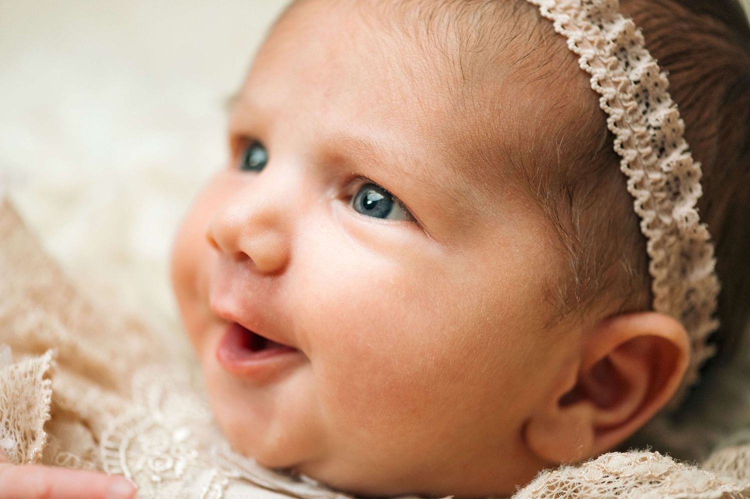 Adorable smiling newborn baby with blue eyes wearing a lace headband, lying on a soft beige blanket.