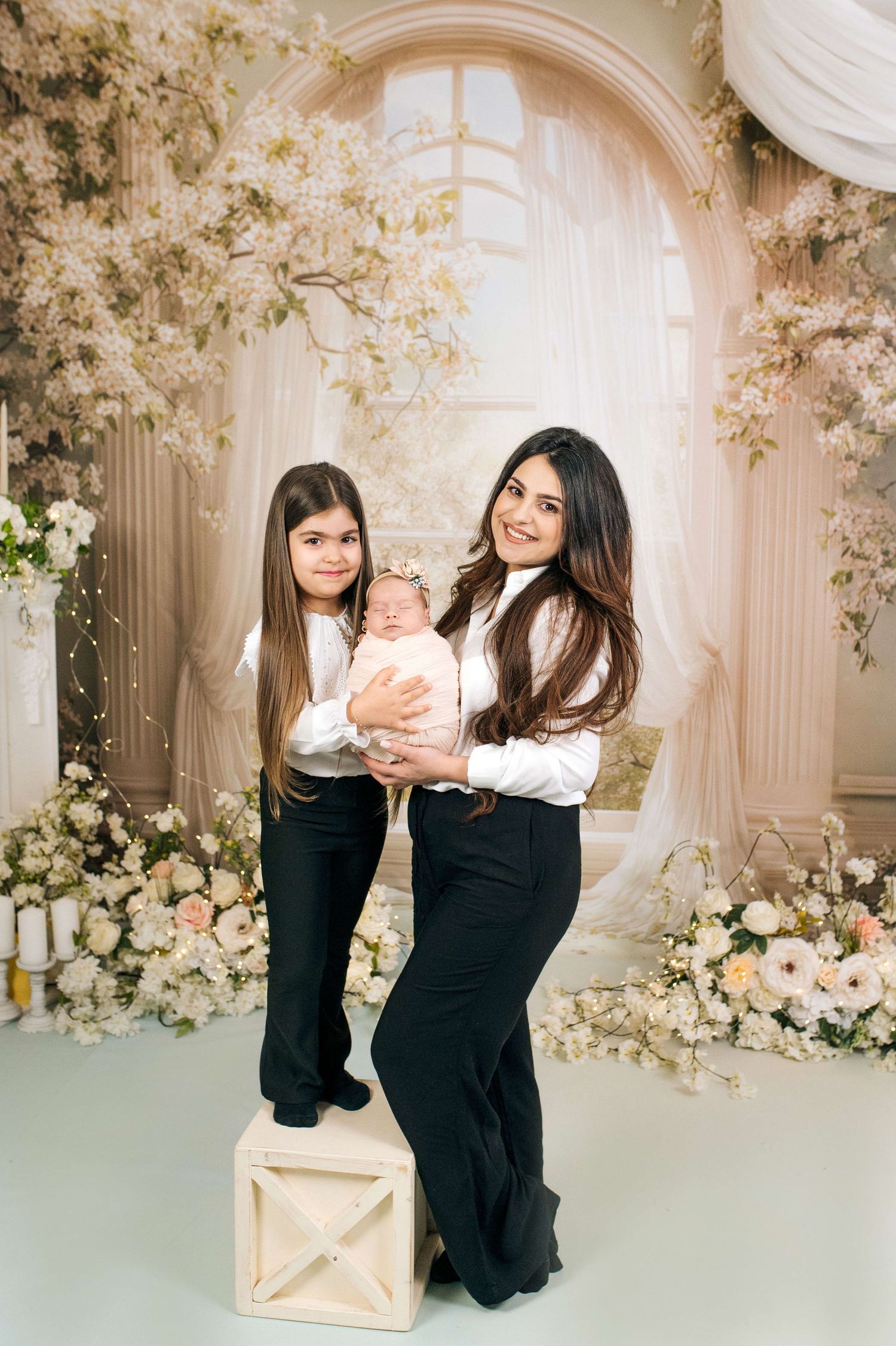A woman holding a newborn and posing with a young girl in matching black and white outfits amid floral decor.