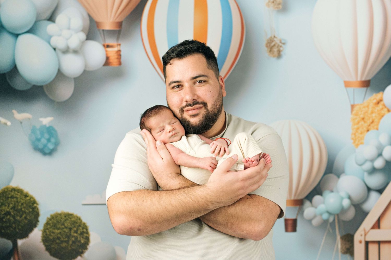 Father holding sleeping newborn baby in front of whimsical hot air balloon and pastel blue balloon backdrop.