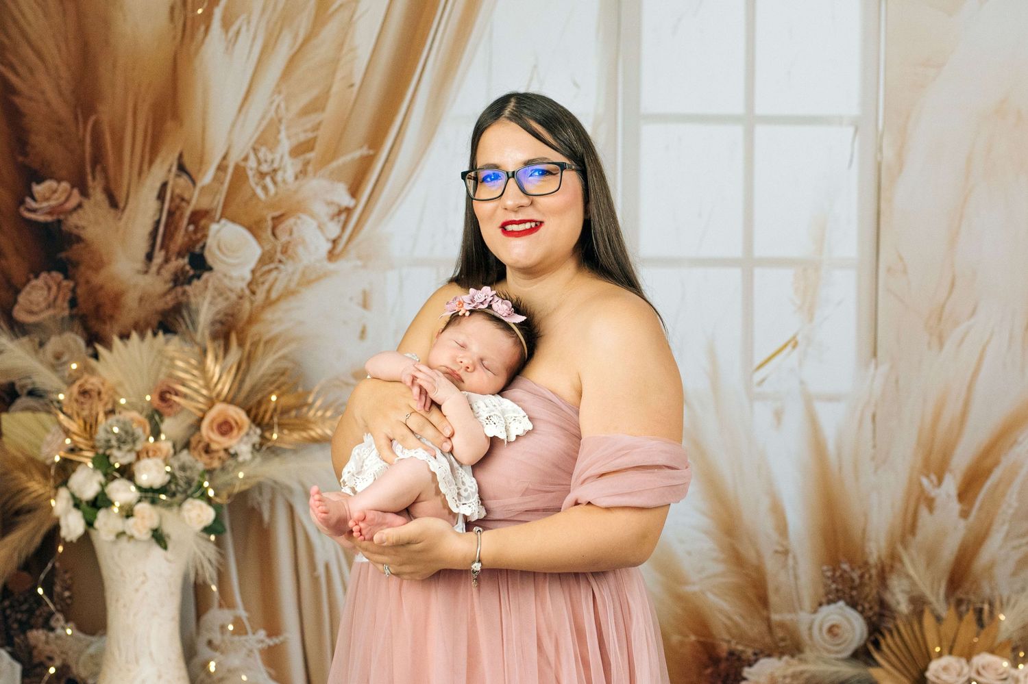 A woman in a pink dress holding a sleeping newborn baby, surrounded by dried floral arrangements in a studio setting.