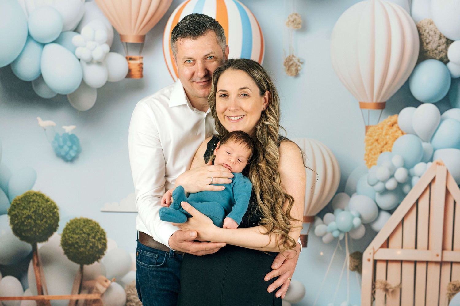 Happy family with newborn baby posing in front of pastel blue balloon and hot air balloon themed backdrop.