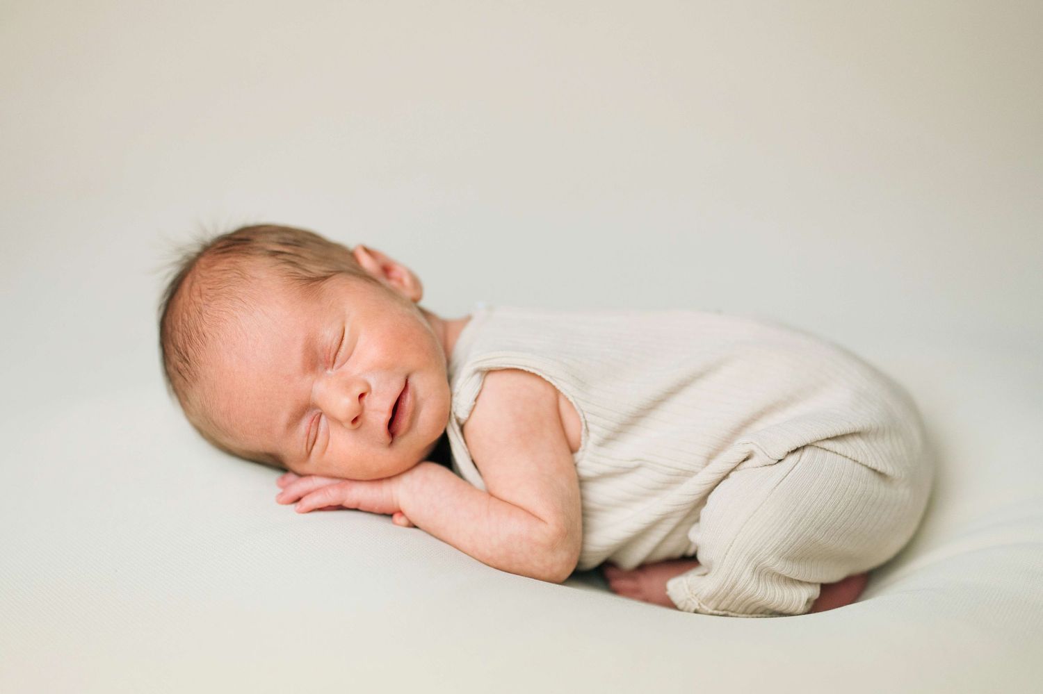 Peaceful newborn baby sleeping curled up on a white blanket wearing a cream outfit in a studio photo.