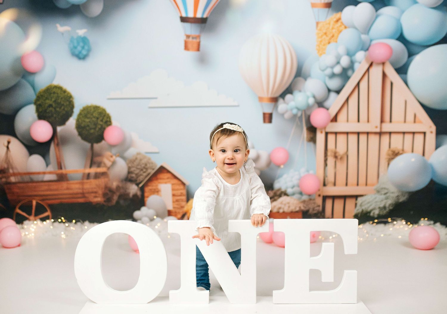 Baby girl celebrating her first birthday with large ONE letters, balloons, and hot air balloon backdrop.