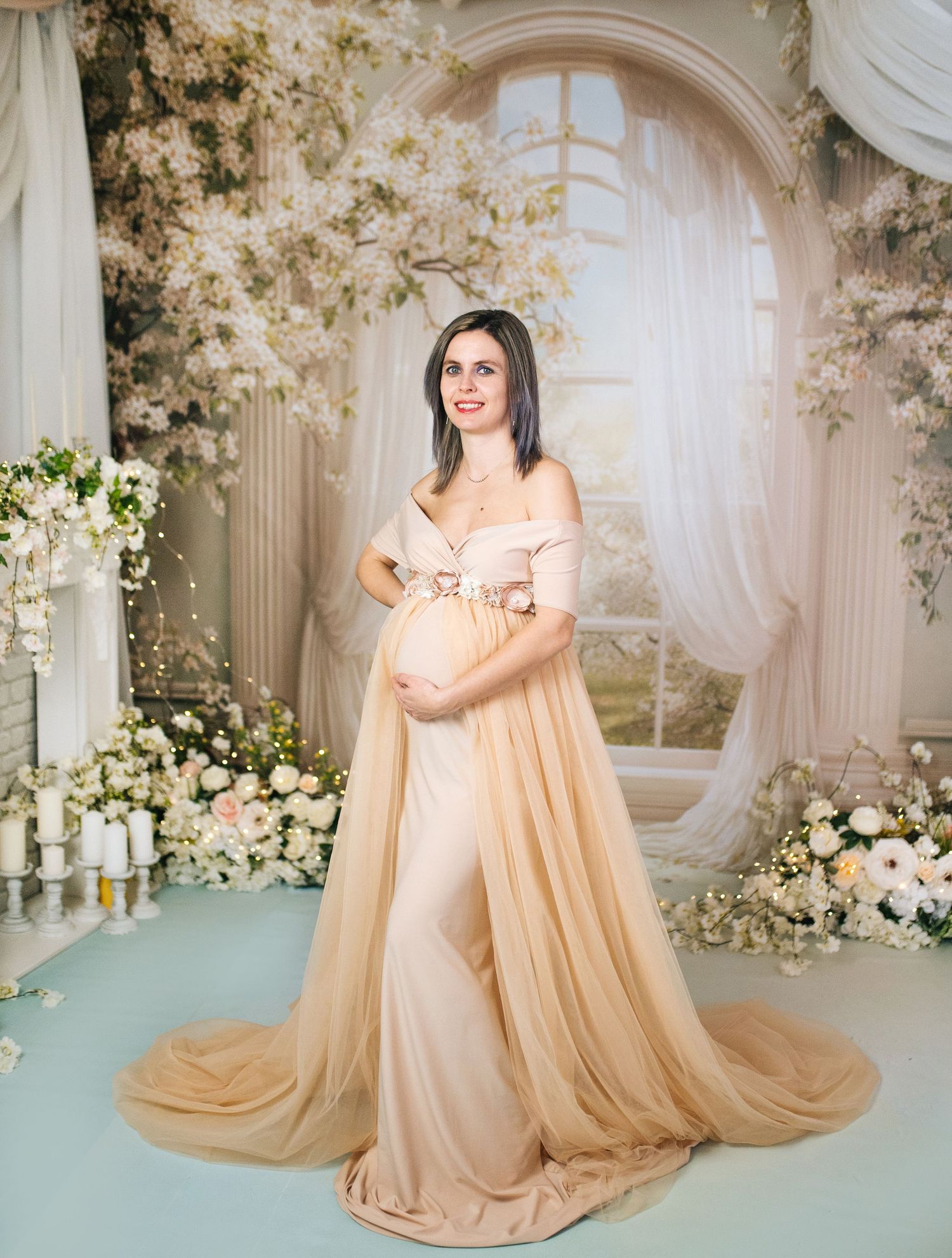 Pregnant woman in elegant gold off-shoulder gown poses gracefully among white floral arrangements in a dreamy studio setting.