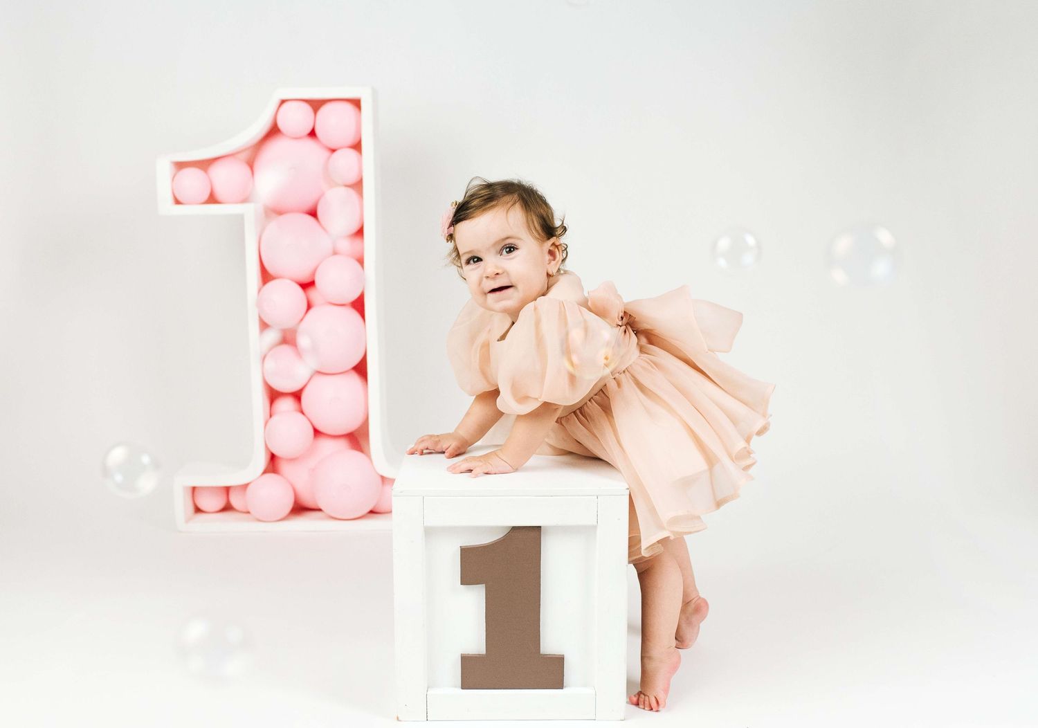 Baby girl in pink dress poses beside a large number 1 balloon display for her first birthday photo shoot.