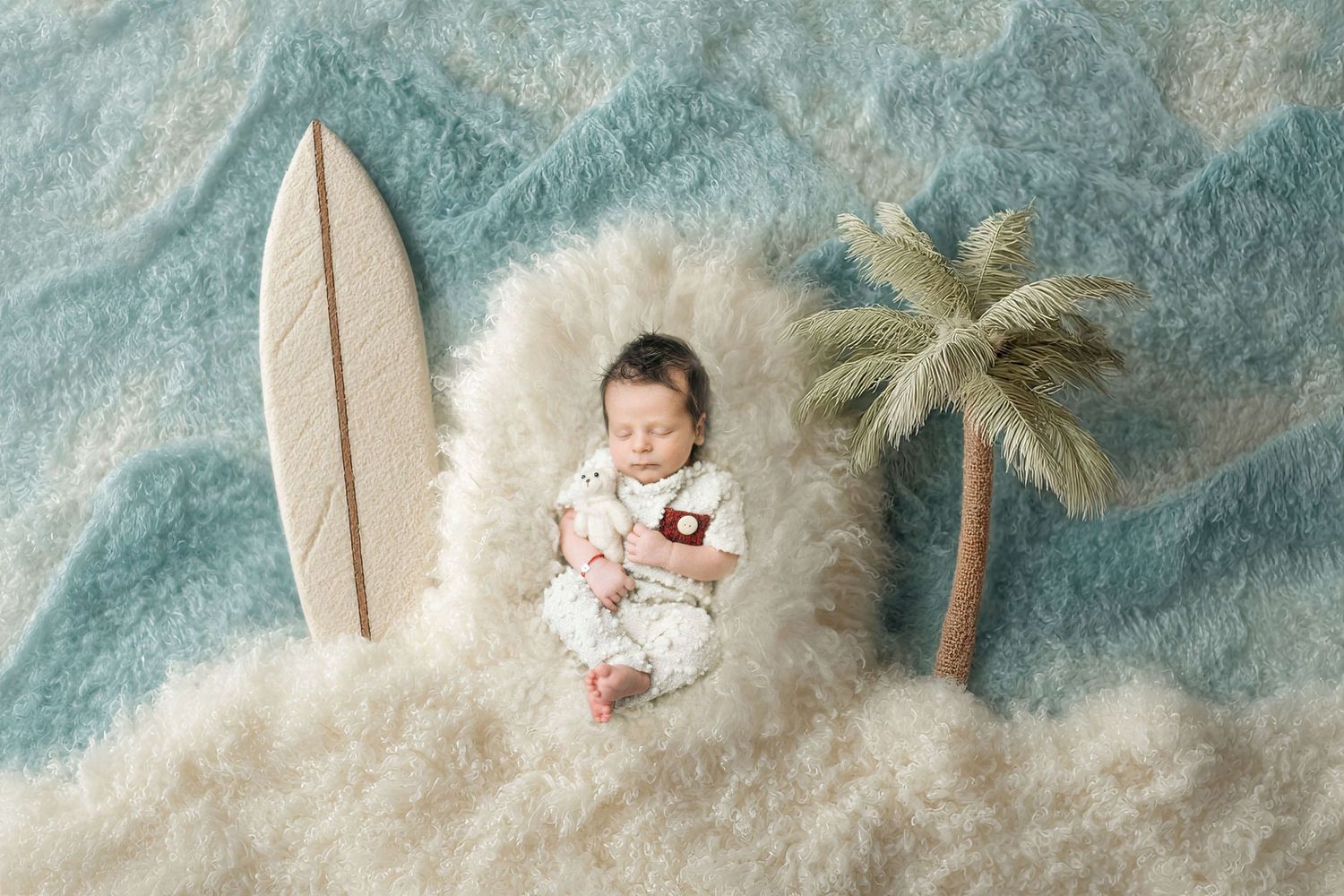Newborn baby sleeping in fluffy white nest with surfboard and palm tree on blue wave backdrop.