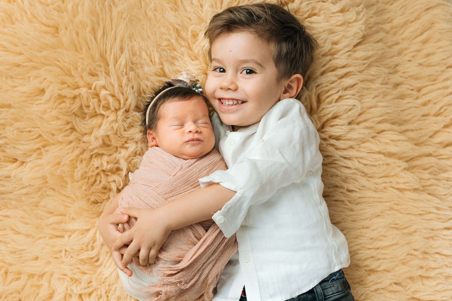 Smiling toddler boy lying on fluffy beige rug hugging swaddled newborn baby sister wearing pink headband.