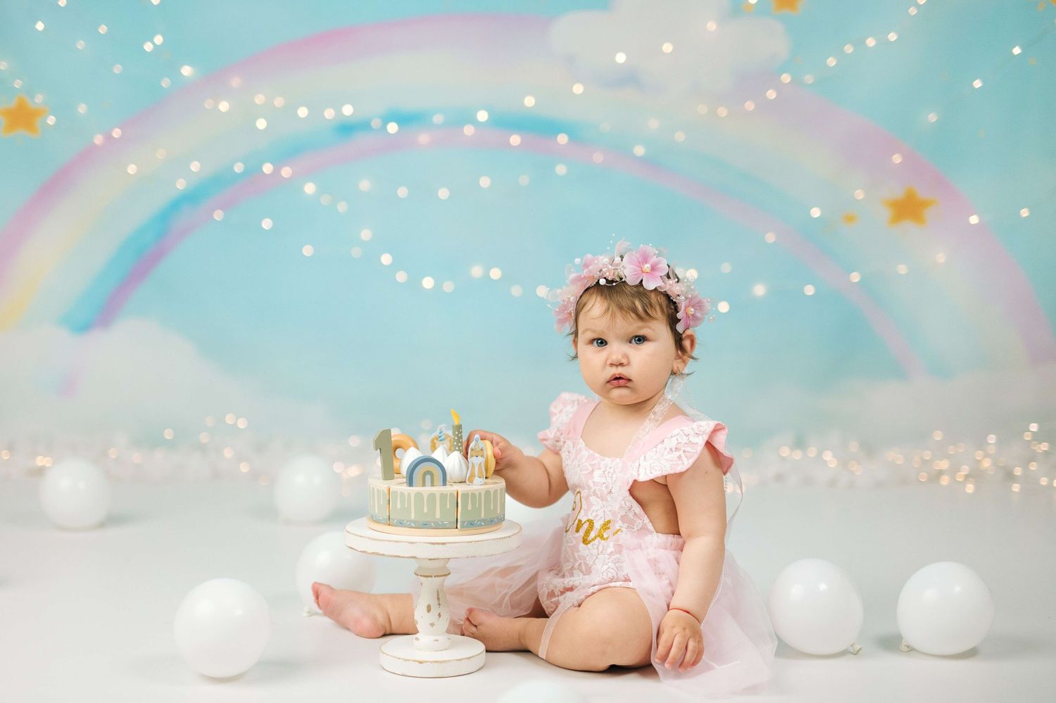 Baby girl in pink romper touches first birthday cake during rainbow-themed cake smash photo session.