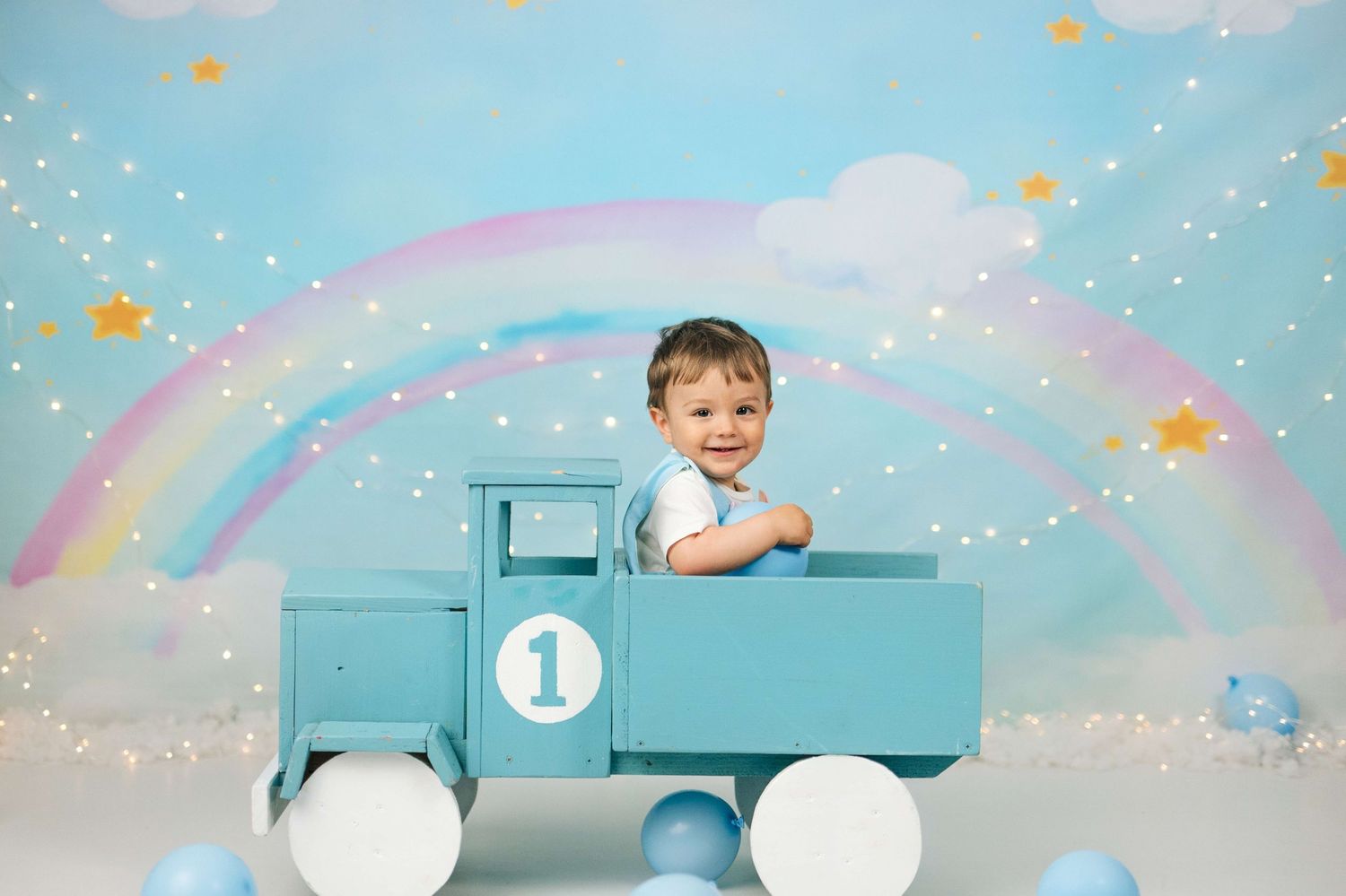 Toddler boy sitting in a blue wooden toy truck with rainbow backdrop and balloons for a first birthday photo.