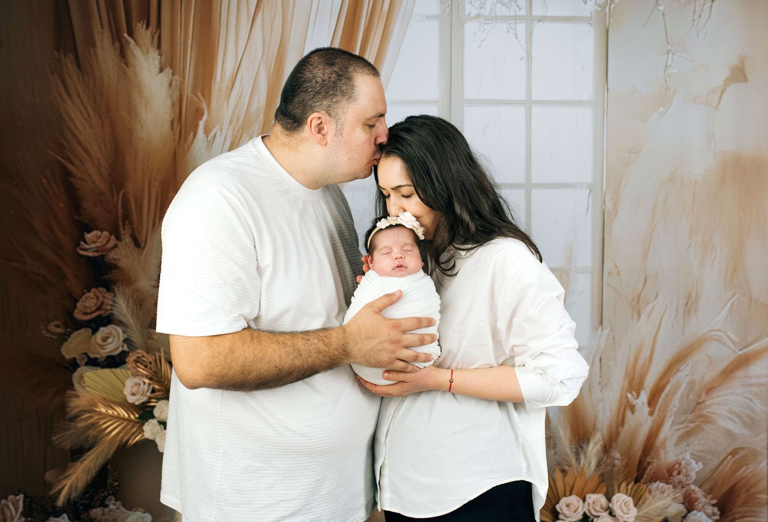 New parents sharing a tender kiss over their swaddled newborn baby during a professional studio photo session.