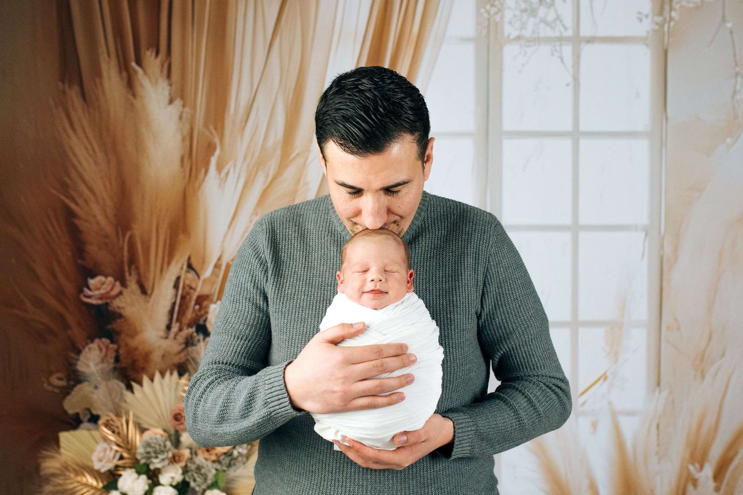 A father tenderly kisses his swaddled newborn baby in a softly decorated studio setting.