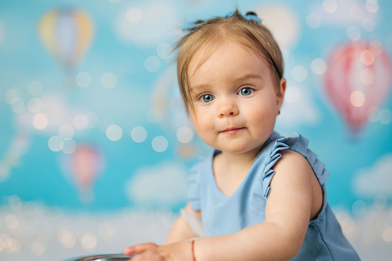 Adorable baby girl in blue dress poses against colorful hot air balloon backdrop with bokeh lighting.
