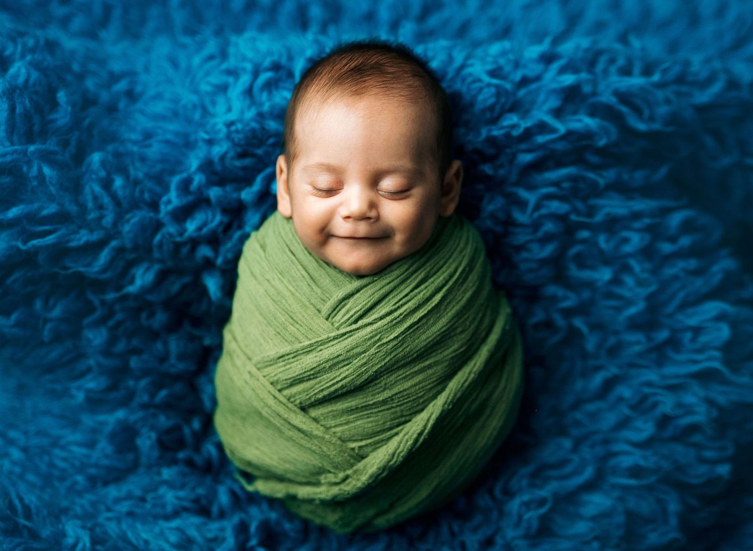 Smiling newborn baby wrapped in green swaddle on blue fluffy blanket during professional newborn photography session.