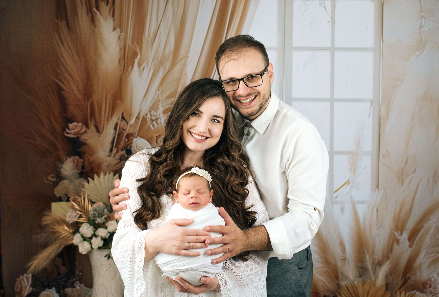 A smiling couple poses with their newborn baby in a boho-styled photo studio with floral decorations.
