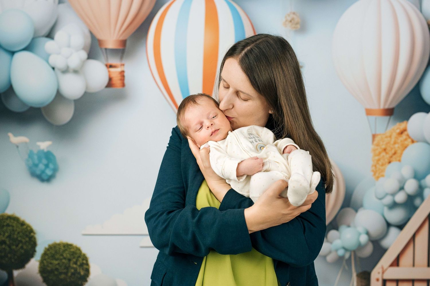 Mother tenderly kissing her sleeping newborn baby against a pastel blue backdrop with hot air balloons.