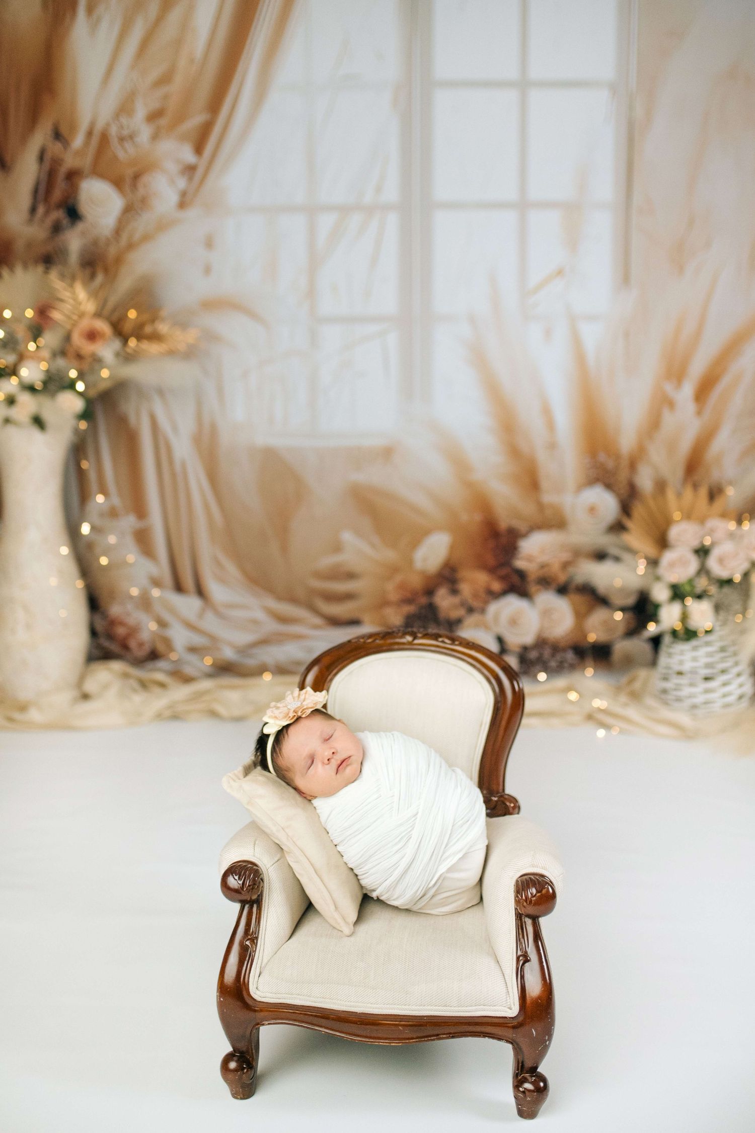 Newborn baby swaddled in white wrap sleeping peacefully on a miniature cream armchair with boho floral backdrop.