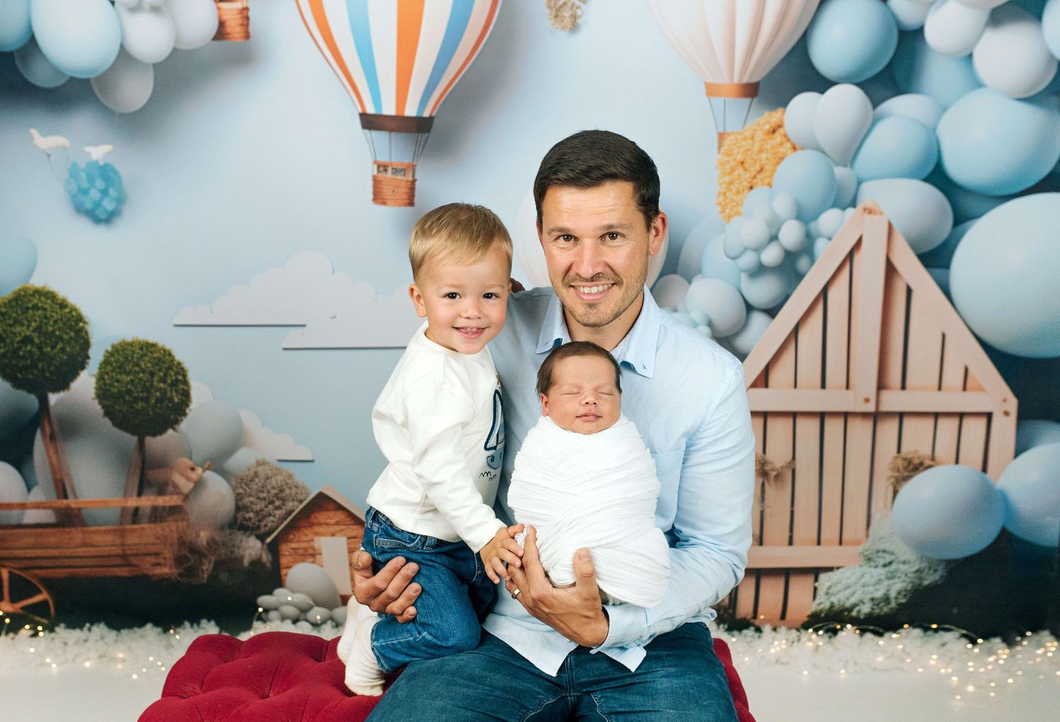 A man sits with two young children in front of a colorful hot air balloon backdrop for a professional photo.