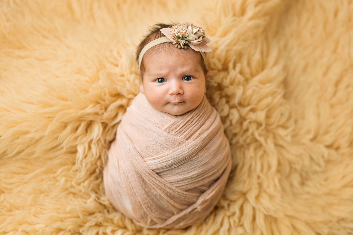 Newborn baby swaddled in pink wrap with floral headband on fluffy yellow fur blanket.