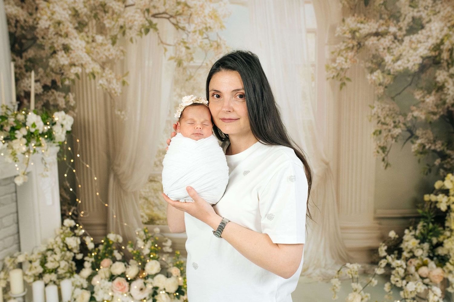 A mother in a white dress tenderly holds her newborn baby, surrounded by elegant floral decorations.