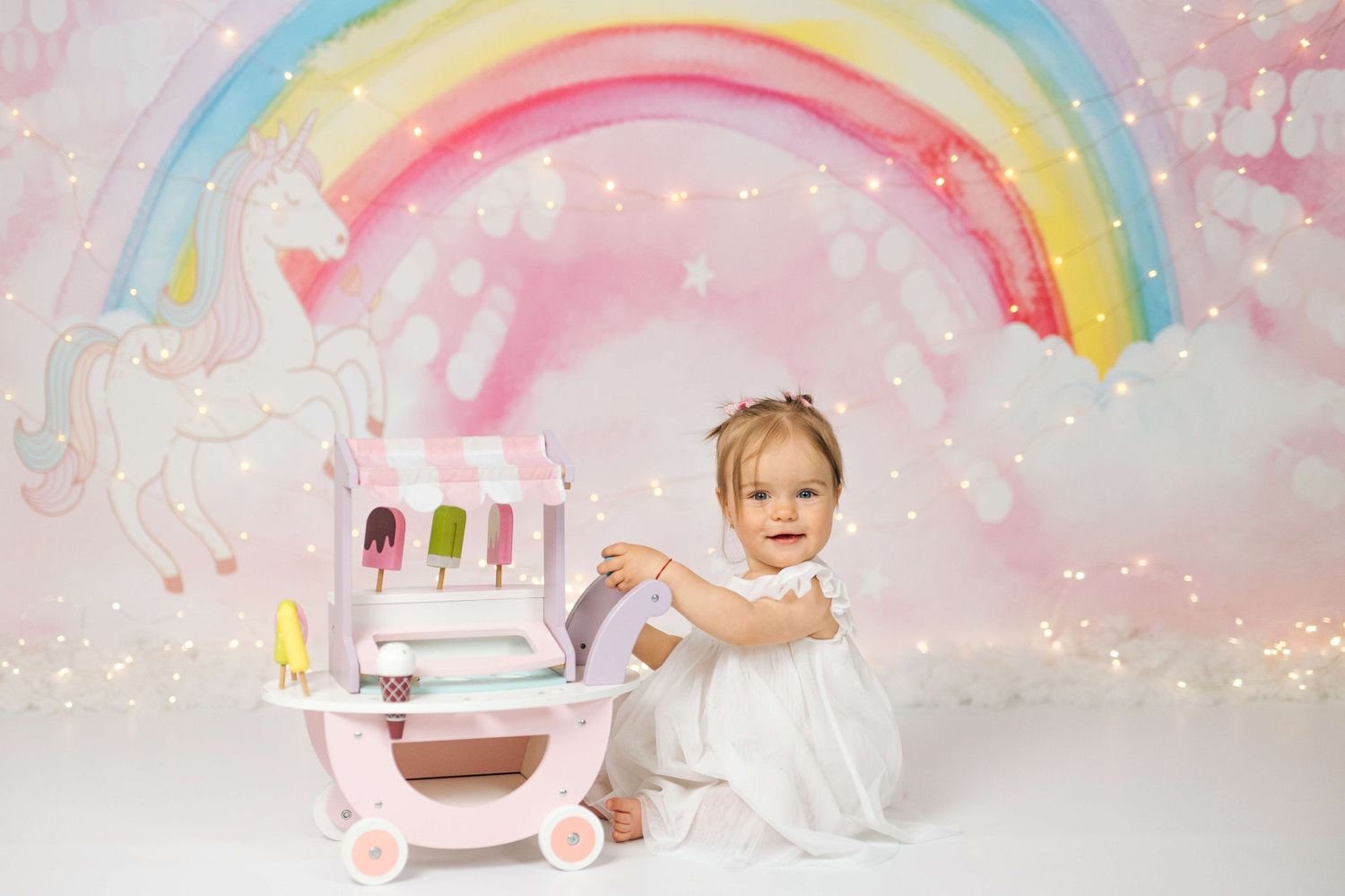 Baby girl in white dress sits beside pink ice cream cart with rainbow unicorn backdrop and fairy lights.
