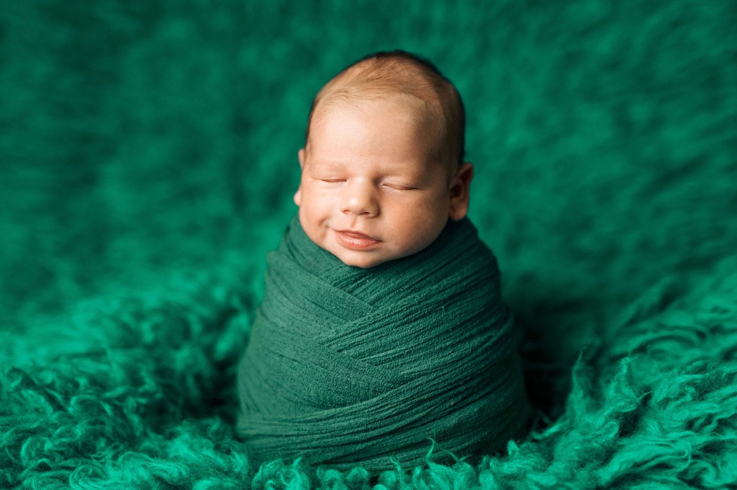 Sleeping newborn baby swaddled in a green wrap, nestled on a soft emerald green fluffy blanket.
