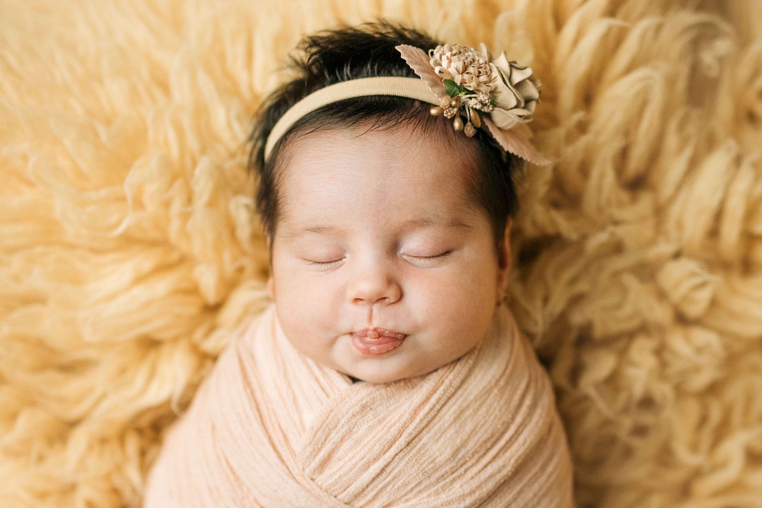 Sleeping newborn baby swaddled in peach wrap with floral headband on fluffy yellow fur backdrop.