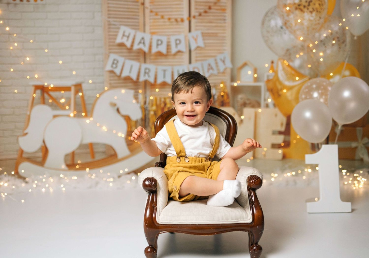 A smiling baby boy in yellow overalls sits in a chair surrounded by balloons and a Happy Birthday banner.