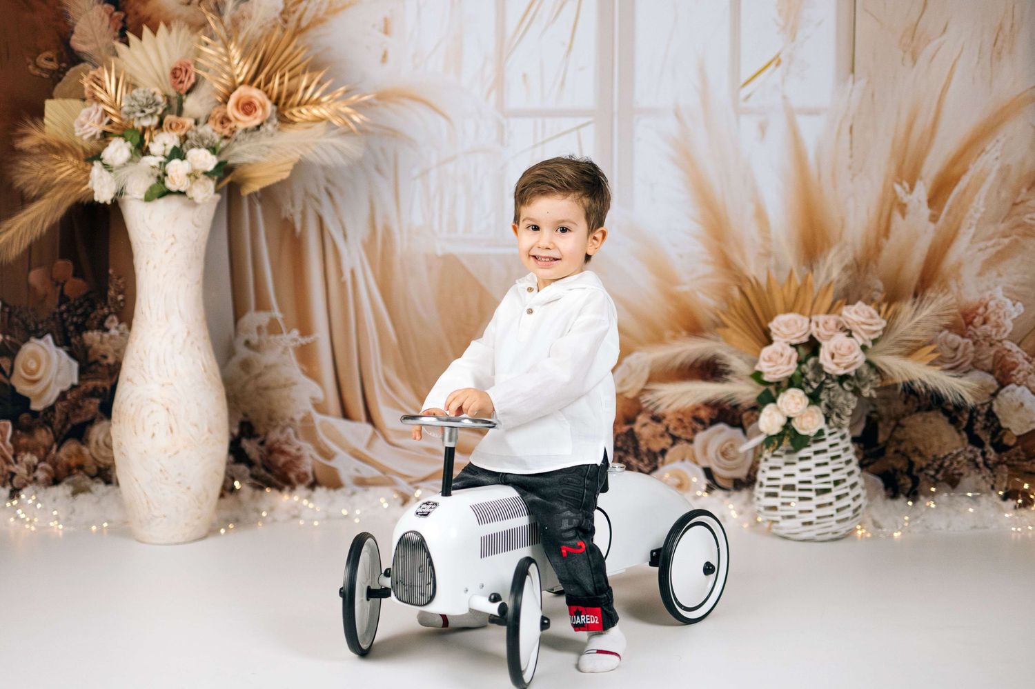 Smiling toddler boy riding a vintage toy pedal car in a boho floral studio photo session.