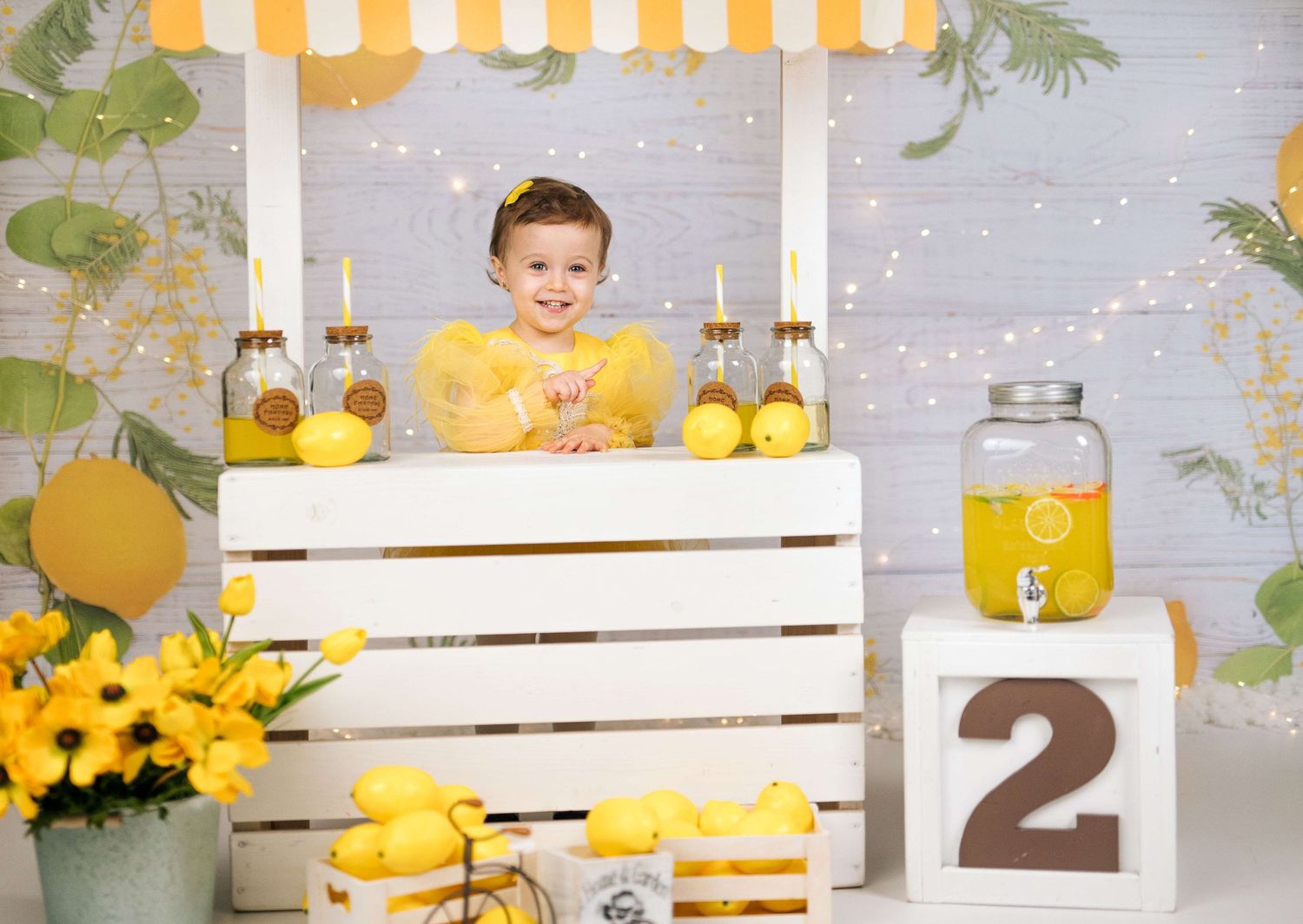 Toddler girl in yellow dress smiling at a lemonade stand with lemons, flowers, and a number 2 block for a birthday photo.