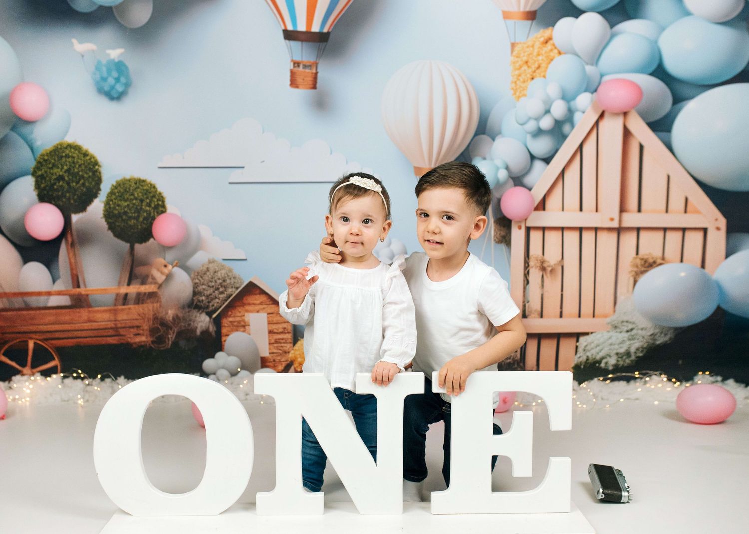 Two siblings smile together beside large white ONE letters in a colorful balloon-themed first birthday portrait.