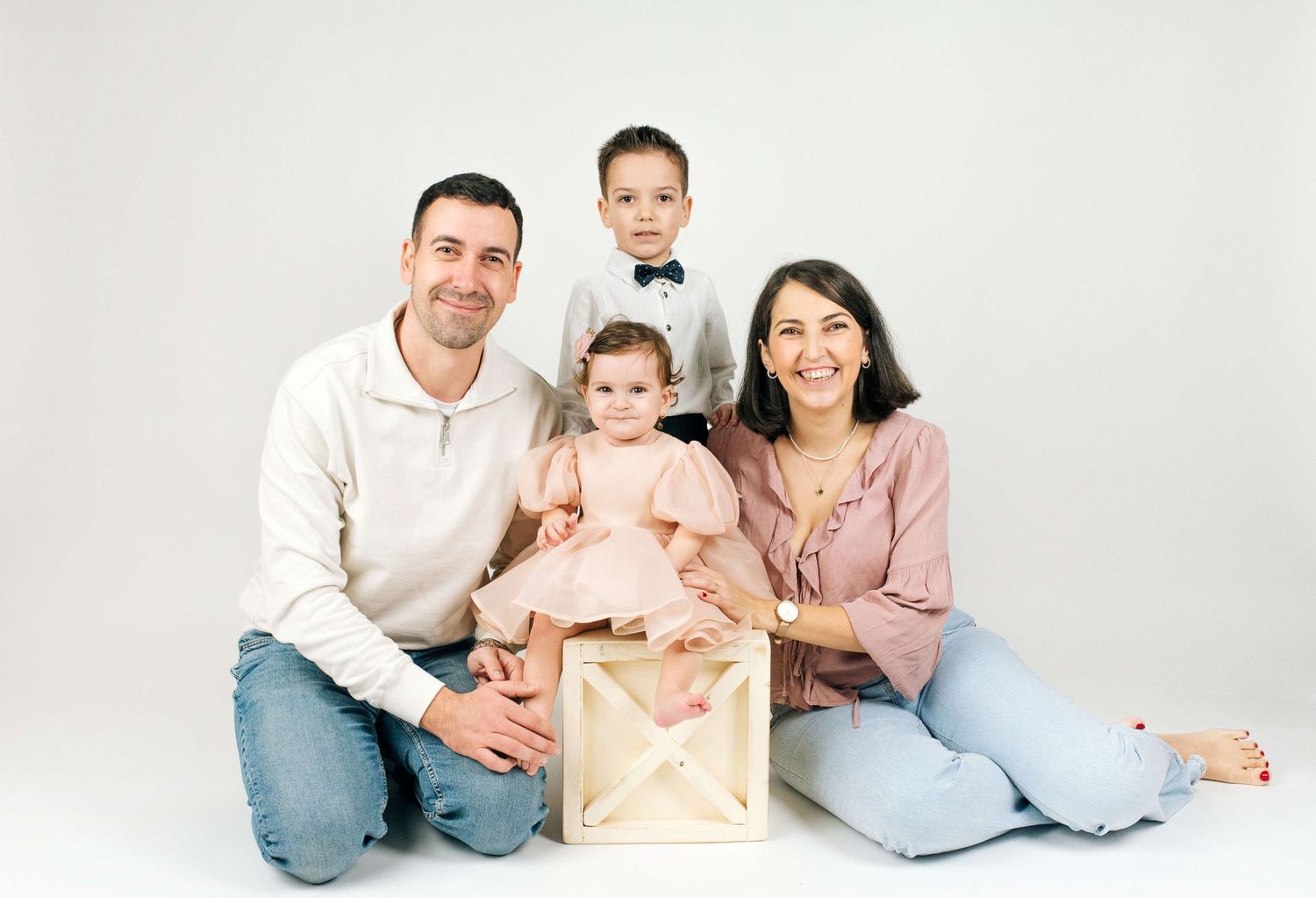 A happy family of four poses in a studio, with parents sitting and two young children, one in a pink dress.