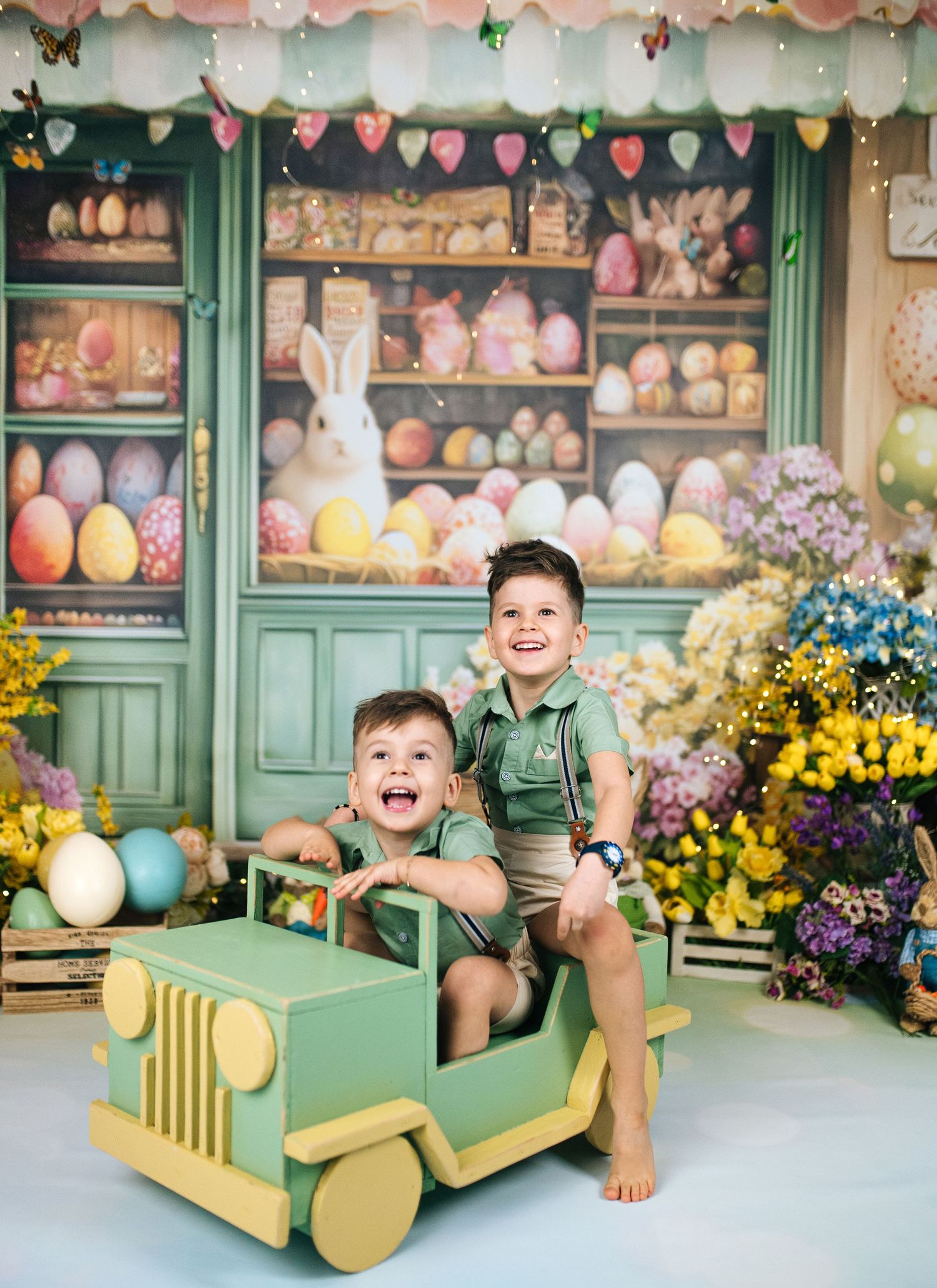 Two smiling boys sit on a toy green Jeep in an Easter-themed photo backdrop with colorful eggs and flowers.
