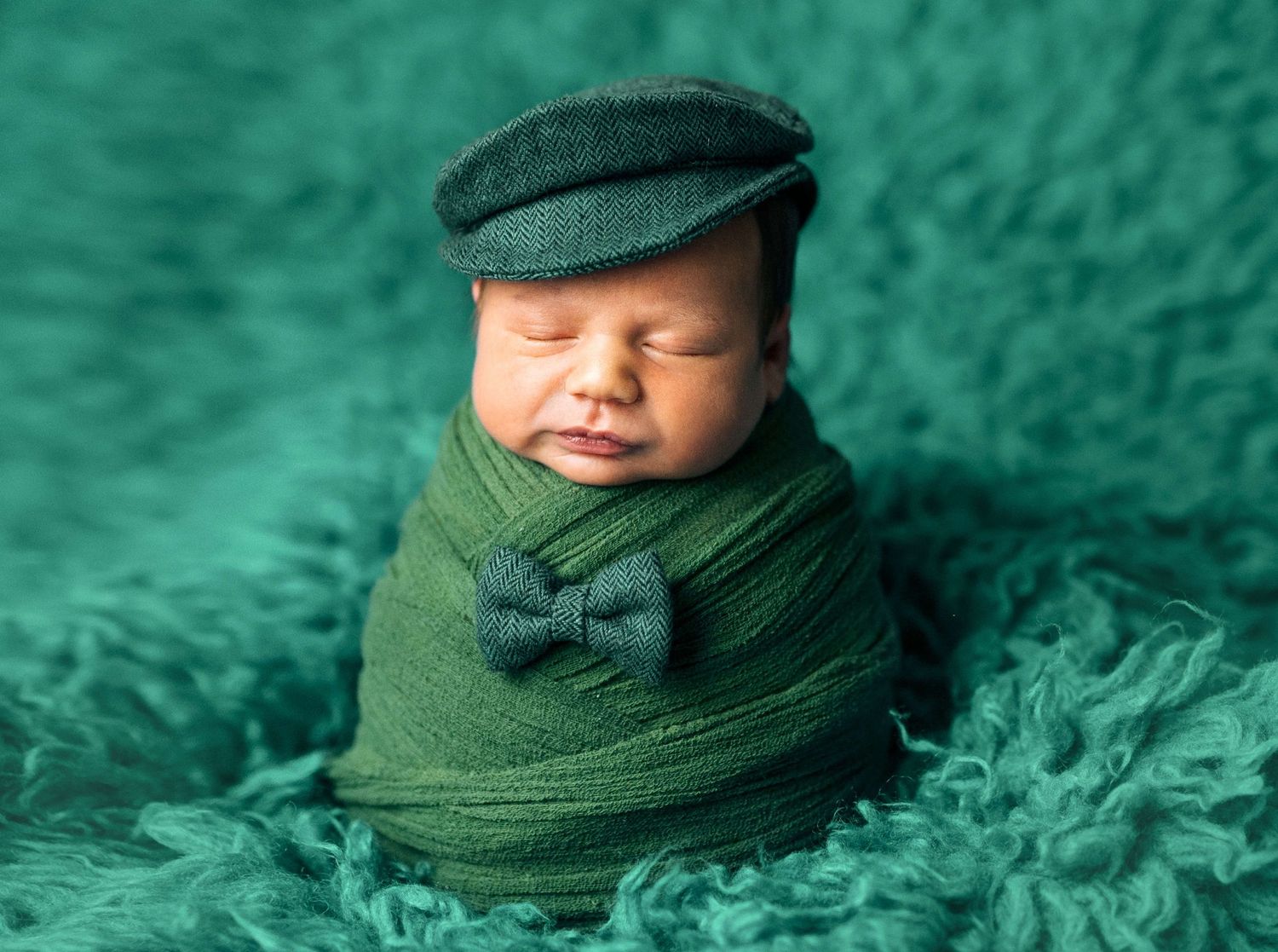 Sleeping newborn baby wrapped in green cloth wearing a flat cap and bow tie on a teal fluffy blanket.