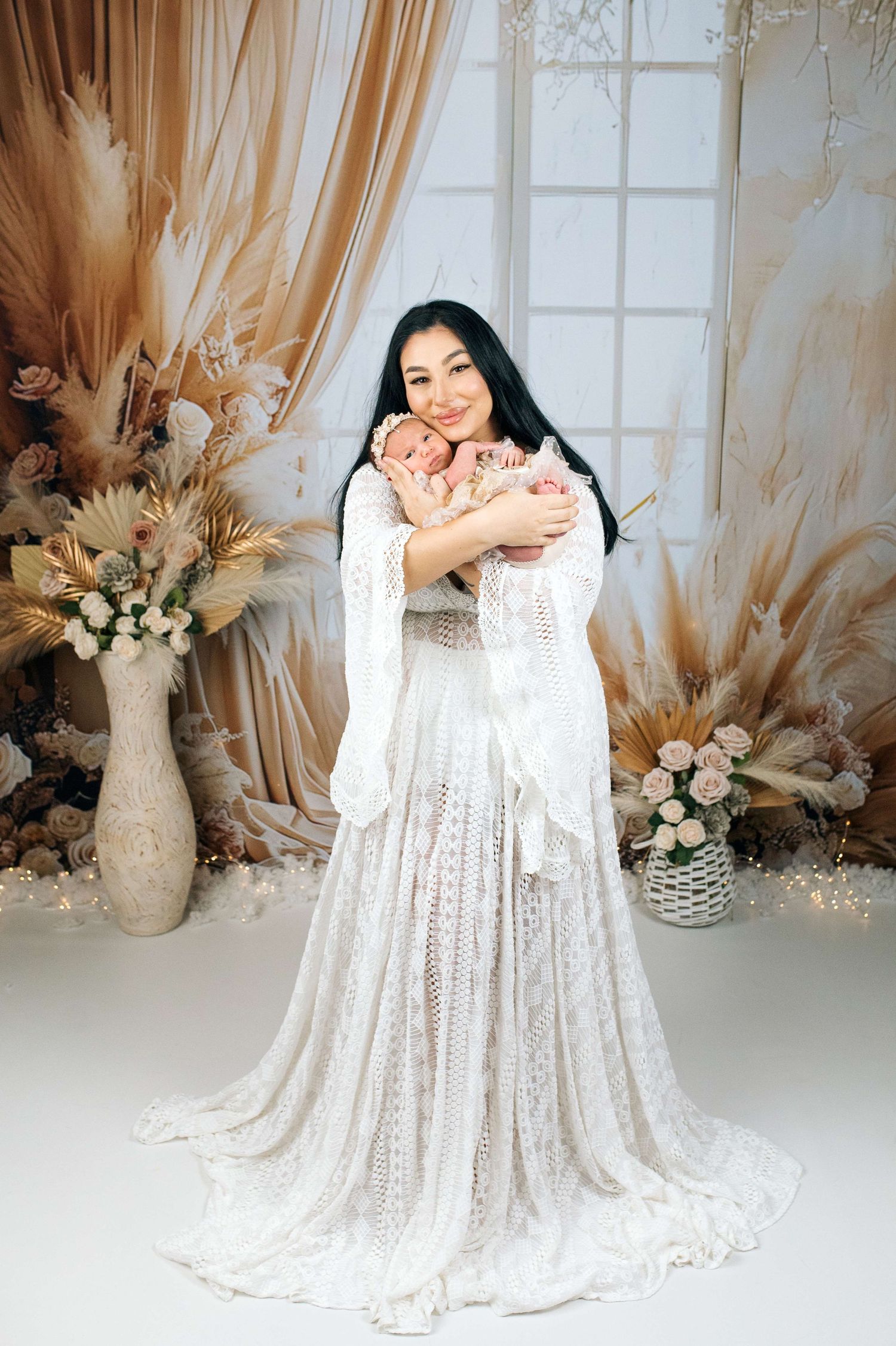 Woman in white lace boho dress holding newborn baby, surrounded by dried floral arrangements in elegant studio setting.