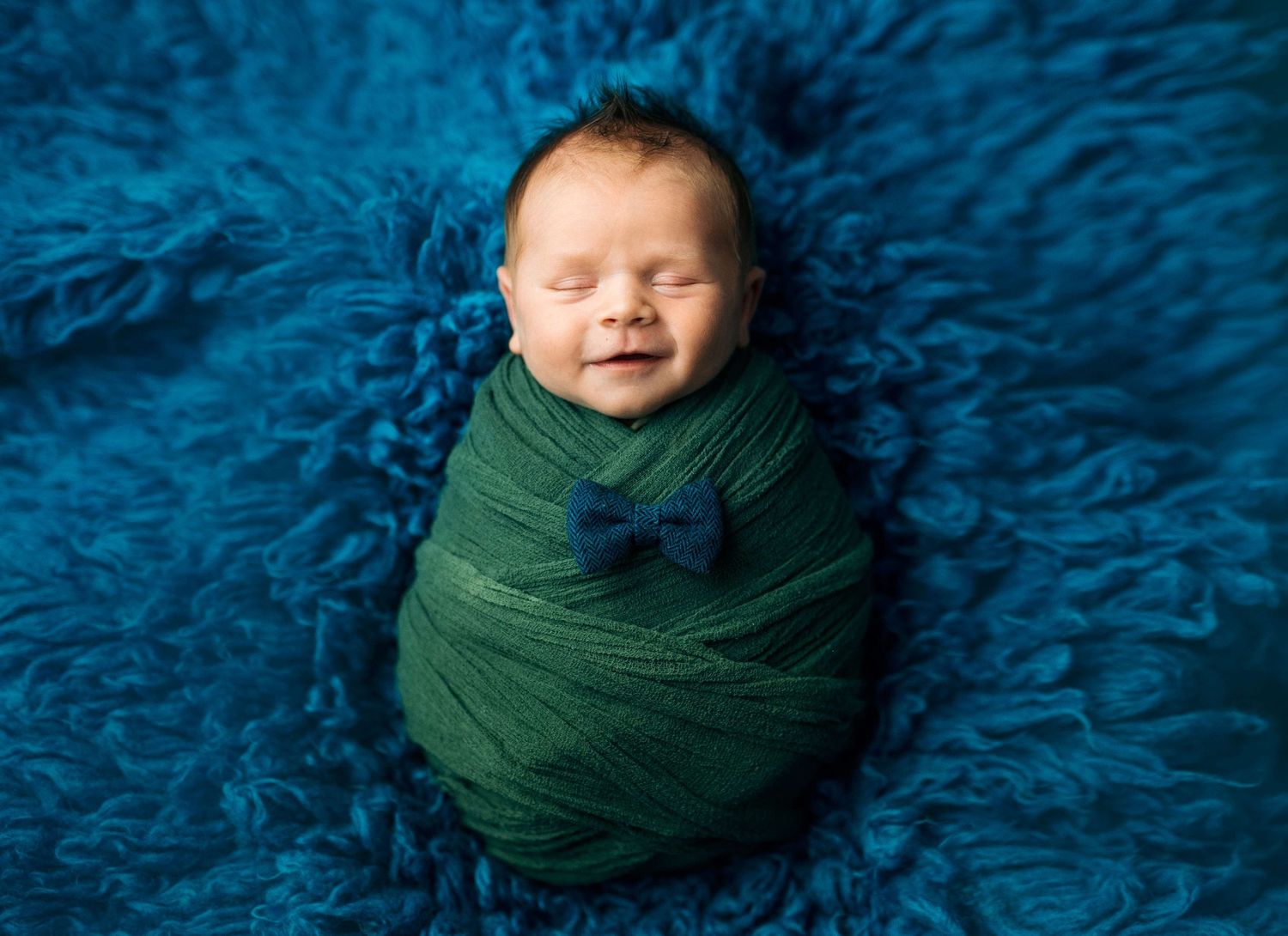 Smiling newborn baby swaddled in green wrap with blue bow tie on fluffy teal rug.