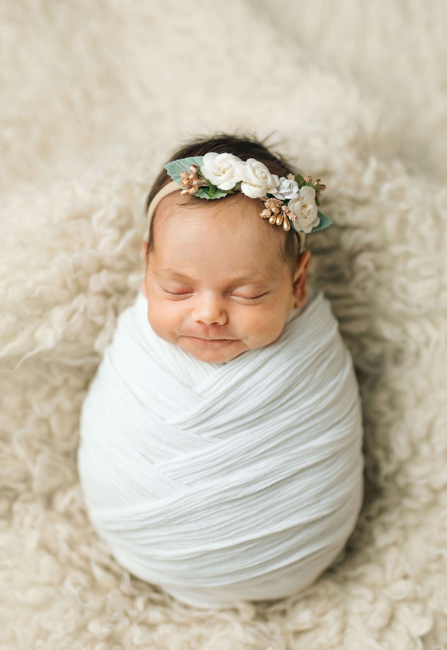 Sleeping newborn baby swaddled in white wrap wearing a delicate floral crown on a cream fluffy blanket.