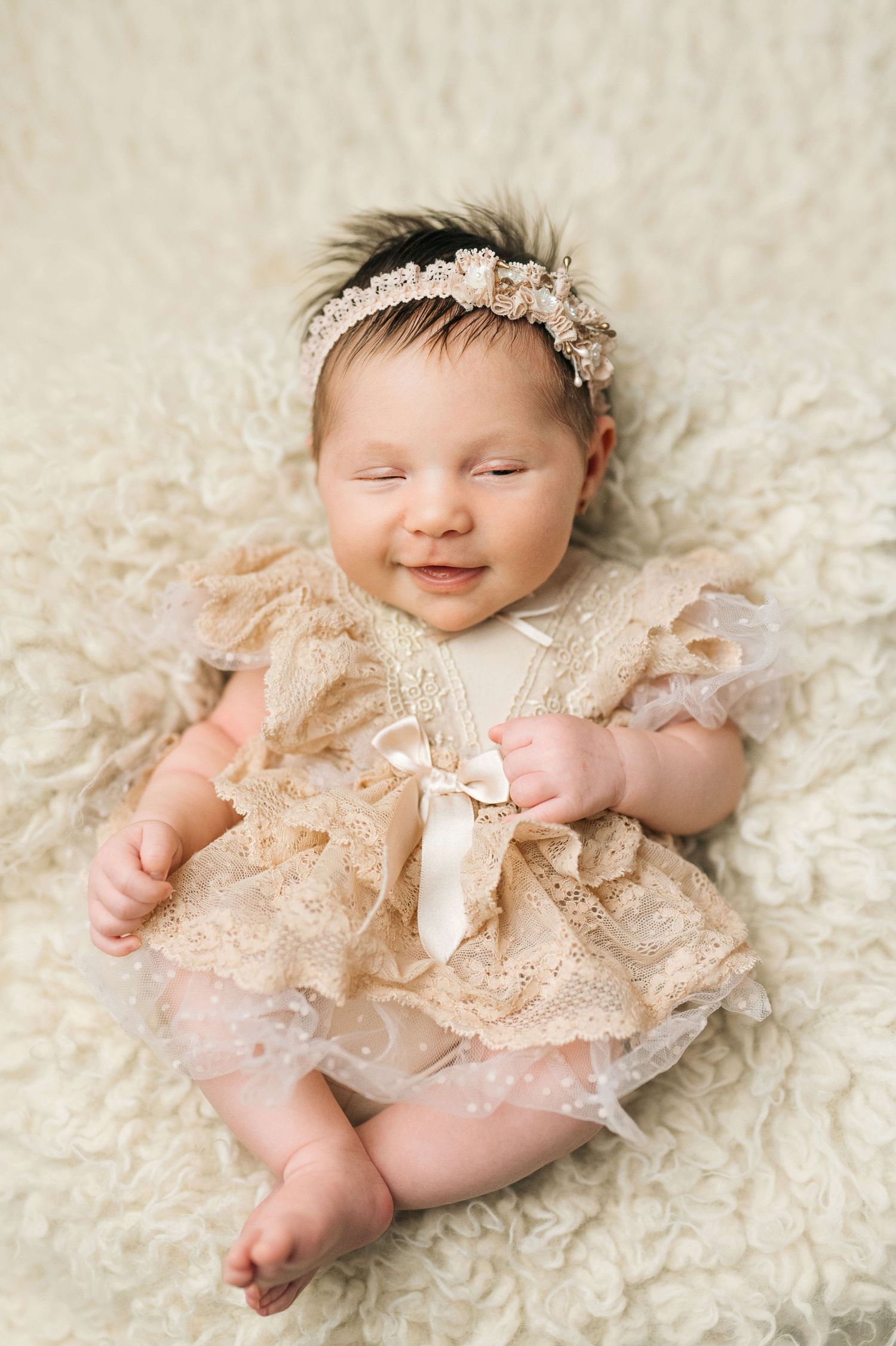 Smiling newborn baby girl in beige lace dress and floral headband lying on fluffy white blanket.