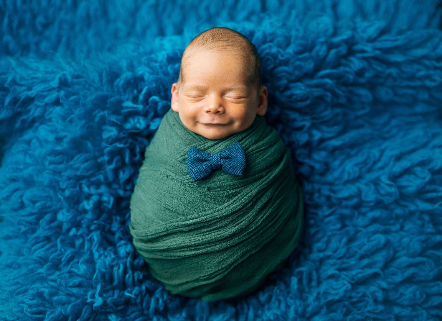 Smiling newborn baby swaddled in green wrap with bow tie on blue fluffy blanket.