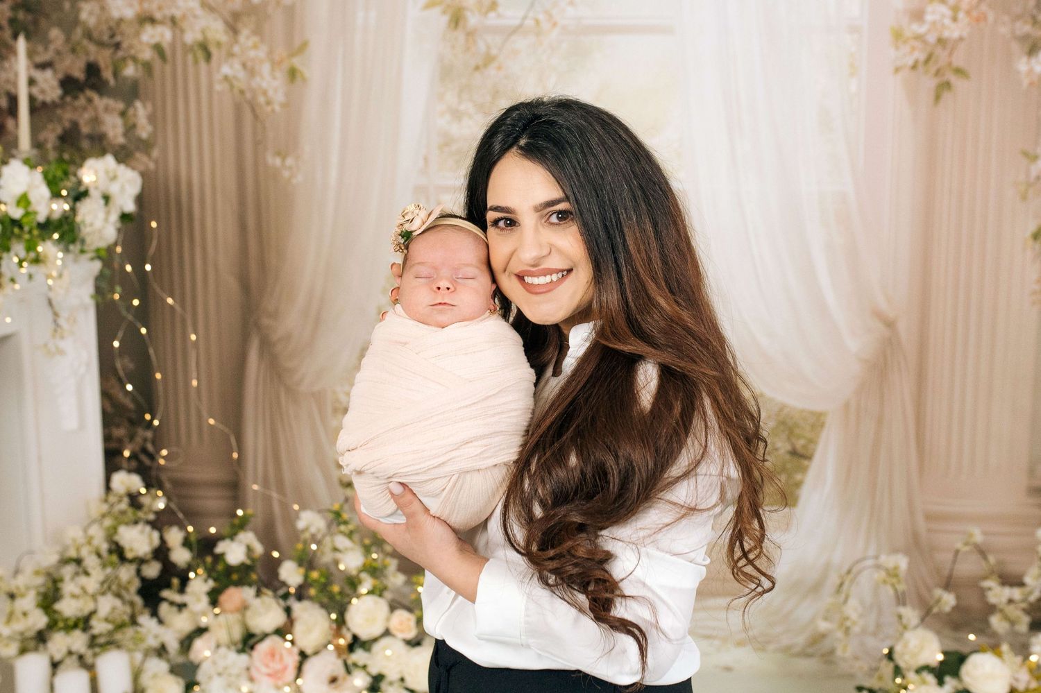 Young woman with long dark hair holds a swaddled newborn baby in a floral decorated studio setting.