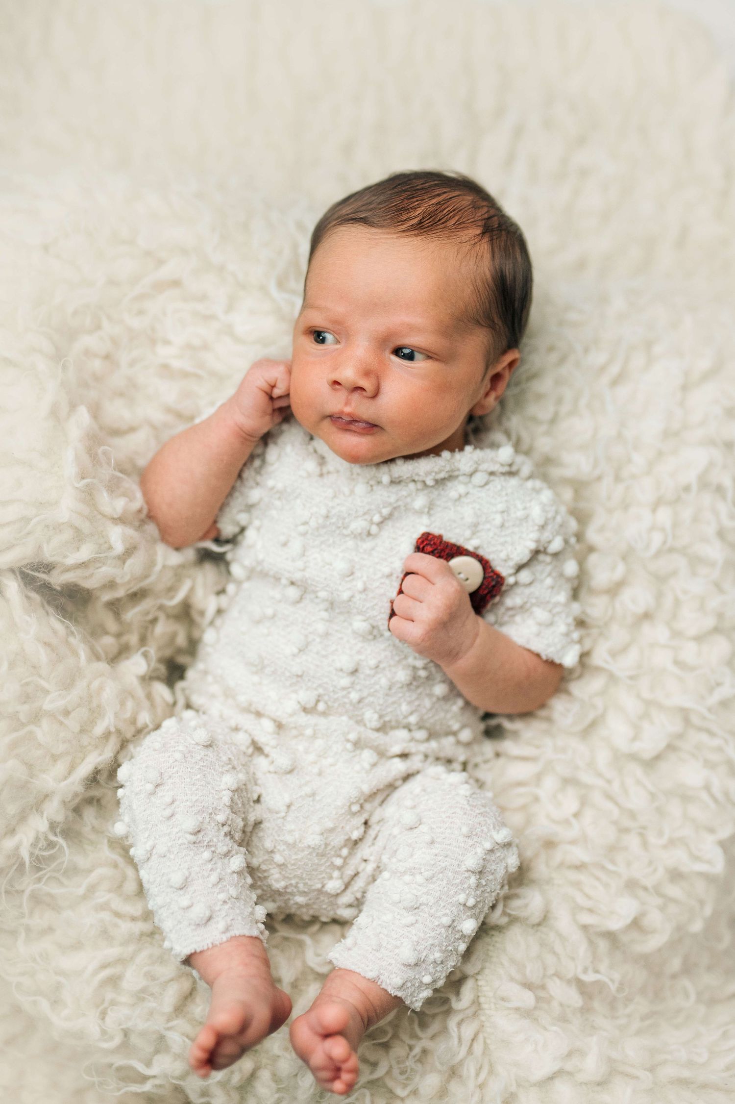 Newborn baby in white textured romper lying on fluffy cream blanket, alert with dark hair and tiny bare feet.